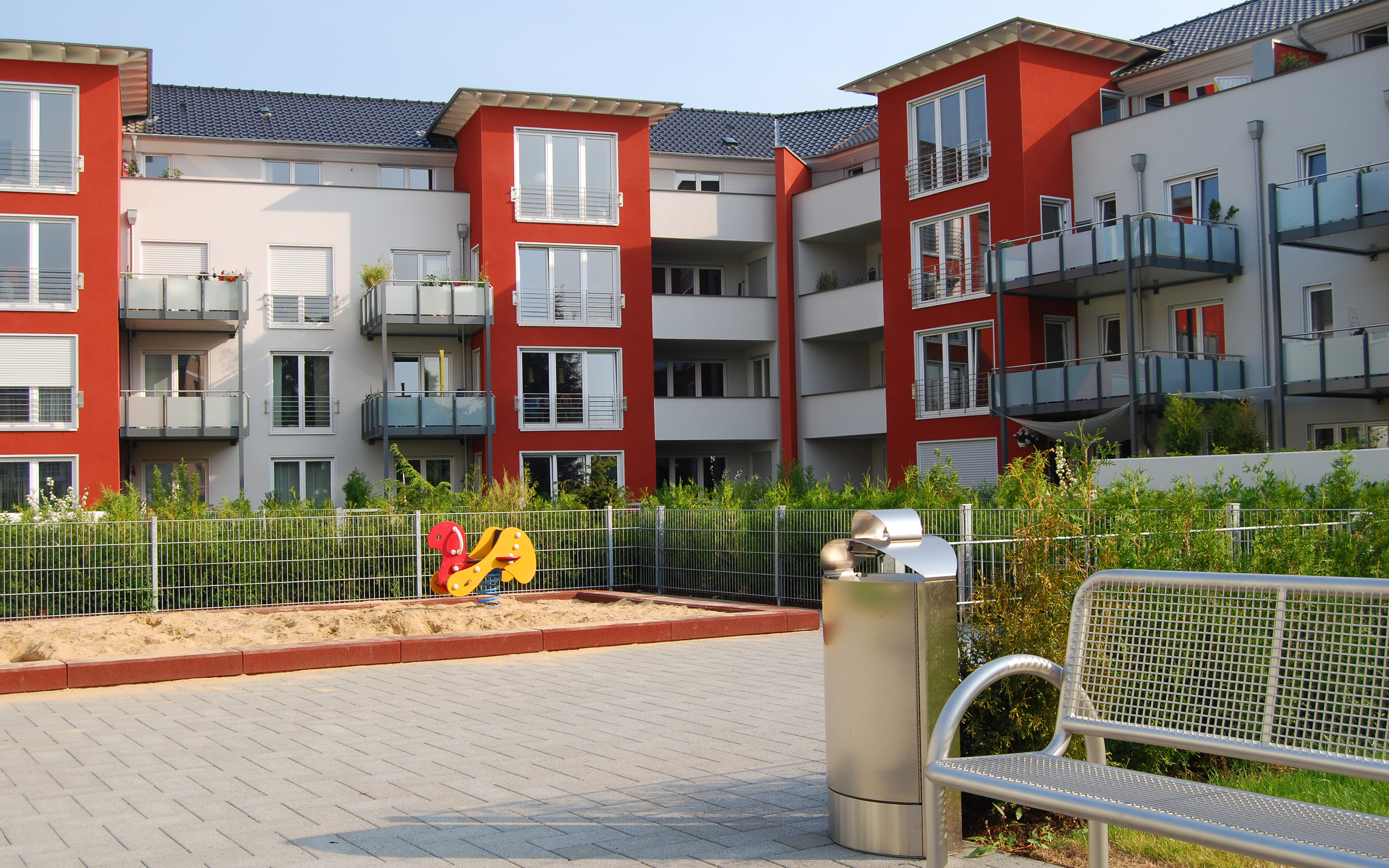 Spielplatz mit Sandkasten und Bank, umgeben von einer Wohnanlage mit rot-weißen Häusern