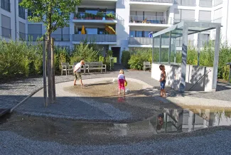 Drei Spring-ins-Feld haben ihre Freude an den drei Springbrunnen – nur der Treppenaufgang erinnert an die Tiefgarage darunter. Spielende Kinder an Springbrunnen und vor dem Treppenaufgang von der Tiefgarage